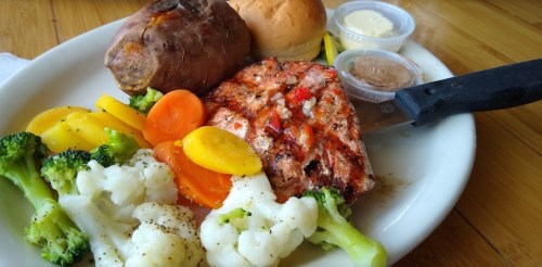 A plate featuring grilled salmon, sweet potato, dinner rolls, and a variety of steamed vegetables.