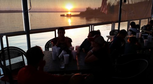 Silhouetted people sitting at a table by the water, watching a sunset over the lake.