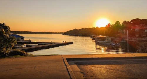 Sunset over a calm lake, with docks and boats lining the shore, surrounded by trees and houses.