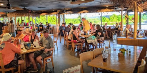 A busy restaurant with patrons dining at tables, tropical decor, and large windows overlooking greenery.