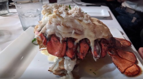 A large lobster tail topped with creamy sauce and crab meat, served on a white plate with garnishes.