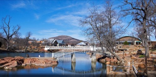 A serene landscape featuring a white bridge over a calm lake, surrounded by trees and distant hills under a clear blue sky.