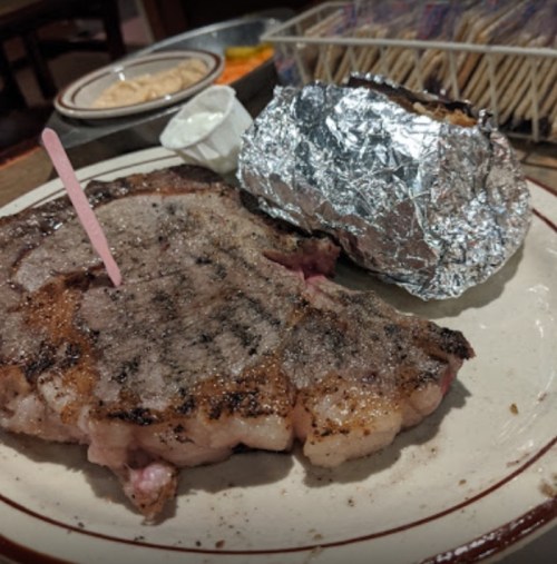 A grilled steak with a pink toothpick and a foil-wrapped baked potato on a plate.