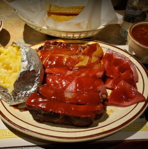 A plate of barbecue ribs with sauce, a baked potato, and a side of beans, served in a casual dining setting.
