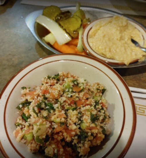 A bowl of tabbouleh salad with tomatoes and herbs, served alongside pickles and hummus with carrot sticks.