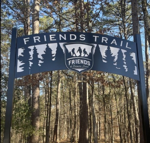 Sign for "Friends Trail" featuring silhouettes of hikers and trees, set in a wooded area.