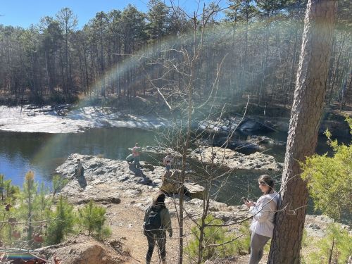 A group of people near a rocky shore by a lake, with a rainbow arching over the water and trees in the background.