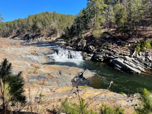 A serene river flows over rocky terrain, surrounded by lush green trees under a clear blue sky.