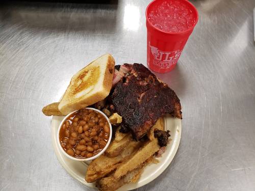 A plate of barbecue ribs, baked beans, toasted bread, and a red cup of drink on a metal surface.