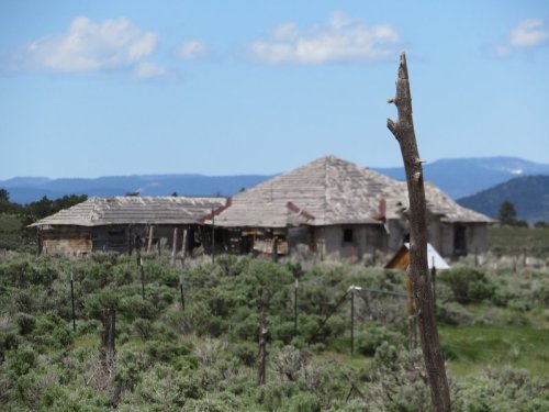 Abandoned rustic house surrounded by sagebrush and mountains under a clear blue sky.