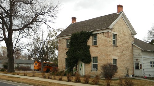 A two-story stone house with ivy on the front, surrounded by bare trees and a grassy area, under a cloudy sky.