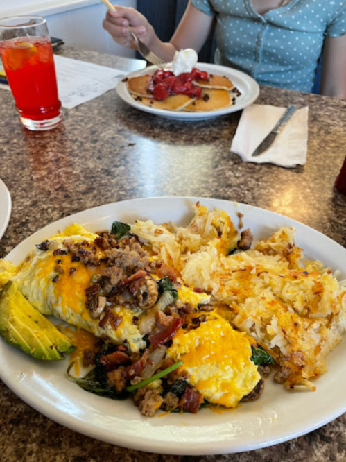 A plate of omelette with cheese, spinach, and bacon, served with hash browns, alongside a person eating pancakes.