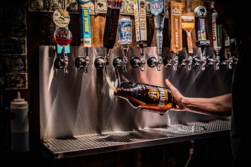 A person fills a growler from a row of beer taps in a bar, with various beer labels visible above the taps.