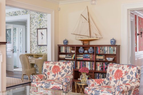 Cozy living room with floral chairs, a bookshelf, and a decorative sailboat on the wall. Bright and inviting atmosphere.