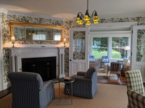 Cozy living room with floral wallpaper, a fireplace, two striped chairs, and a view of a sunlit porch.