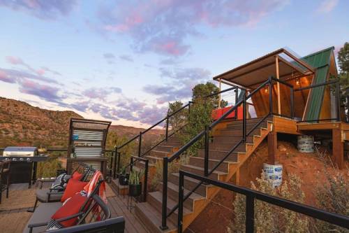 A cozy outdoor deck with seating, leading to a modern cabin on a hillside, surrounded by scenic mountains and a colorful sky.