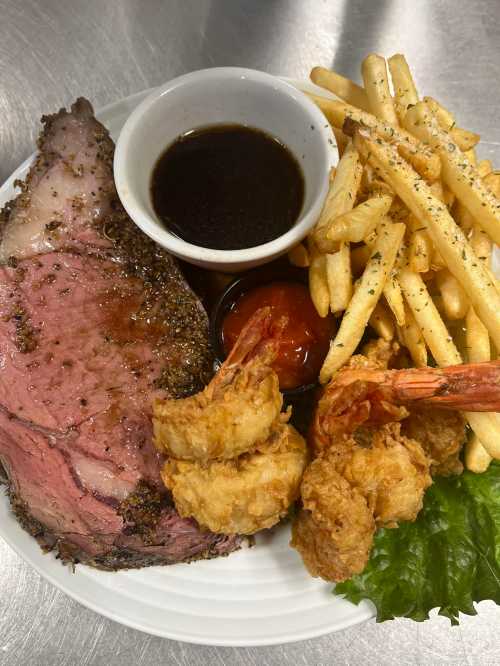 A plate featuring prime rib, fried shrimp, French fries, and dipping sauces on a bed of lettuce.