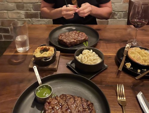 A table setting with two plates of steak, macaroni, and a small dish, accompanied by a glass of wine and water.