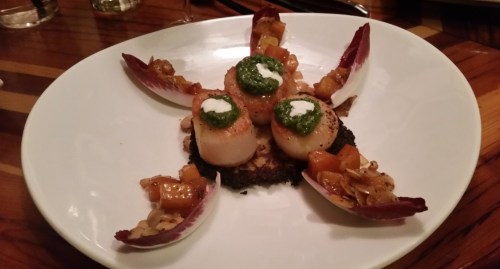 A beautifully plated dish featuring seared scallops with pesto, served on a toasted base and garnished with vegetables.