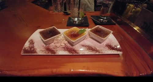 Three small bowls of crème brûlée on a white plate, garnished with whipped cream and a lime slice, on a wooden table.