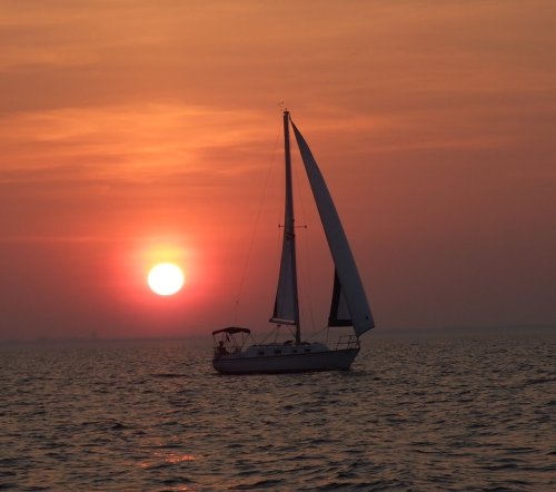 A sailboat glides across the water at sunset, with a vibrant orange sun setting on the horizon.