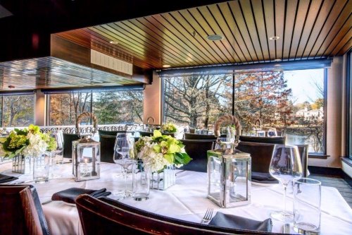 Elegant dining setup with floral centerpieces, glassware, and large windows showcasing a scenic view outside.