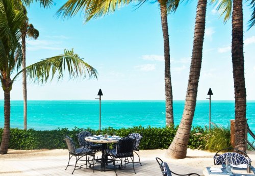 A serene beach view with palm trees, a table set for dining, and turquoise waters under a clear blue sky.