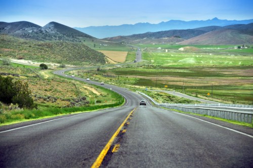 A winding road stretches through green fields and mountains under a clear blue sky.