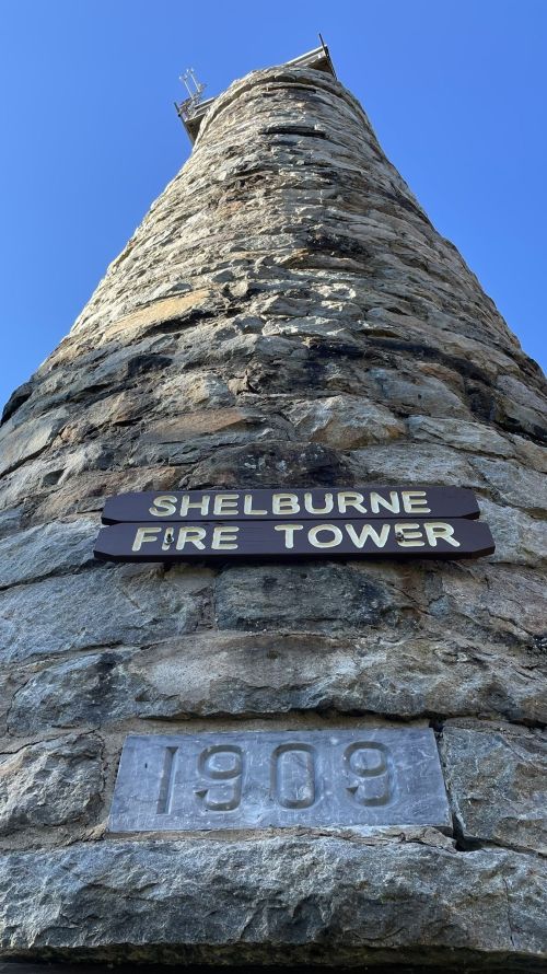 A tall stone fire tower with a sign reading "Shelburne Fire Tower" and the year "1903" visible at its base.