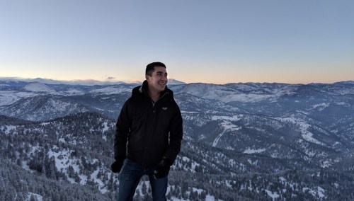 A person stands on a snowy mountain peak, smiling with a scenic view of hills and valleys in the background at sunset.
