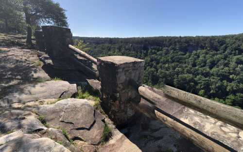 A stone and wooden railing overlooks a lush green valley under a clear blue sky.