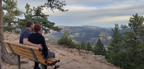 A couple sits on a bench overlooking a scenic mountain view, surrounded by trees and a cloudy sky.