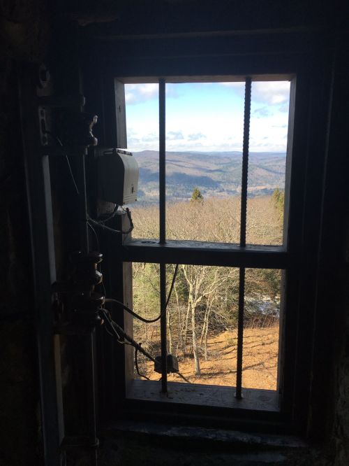 View from a window with a metal frame, showcasing a scenic landscape of trees and mountains under a blue sky.