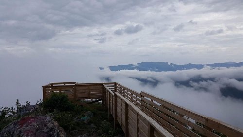 A wooden viewing platform overlooks a sea of clouds and distant mountains under a cloudy sky.