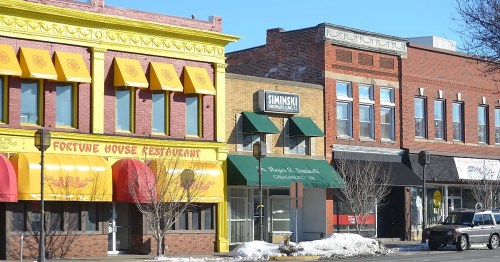 Colorful storefronts line a street, featuring a restaurant and various shops under a clear blue sky. Snow is visible on the ground.
