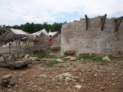 A stone structure with a person near it, surrounded by tents and rocky terrain under a cloudy sky.