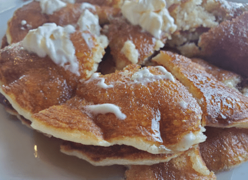 A close-up of golden pancakes drizzled with syrup and topped with dollops of whipped cream on a white plate.