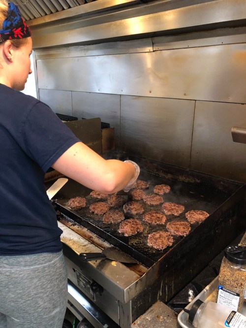 A person cooks multiple burger patties on a griddle in a commercial kitchen. Steam rises from the cooking surface.