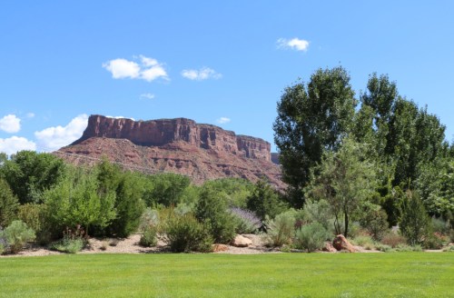 A scenic view of a red rock formation under a blue sky, surrounded by green trees and shrubs in a grassy area.