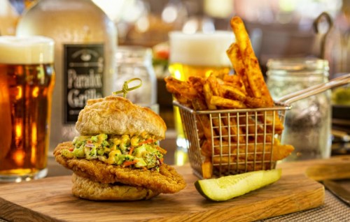 A crispy chicken sandwich with avocado spread, served with a side of fries in a basket and a pickle slice.