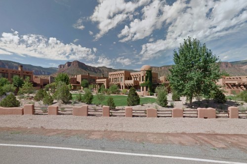 A scenic view of adobe-style buildings surrounded by greenery and mountains under a partly cloudy sky.