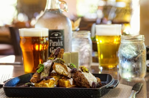 A plate of fried chicken on waffles, topped with sauce and garnished, with two glasses of beer in the background.