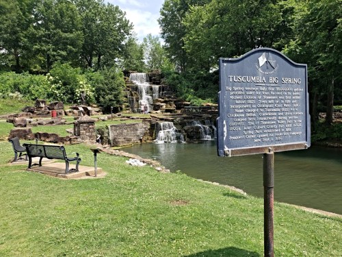 A scenic view of Tuscumbia Big Spring with a waterfall, historical sign, and benches in a lush green park.