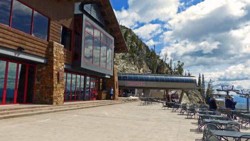 A mountain lodge with large windows, outdoor seating, and a ski lift in the background under a cloudy sky.