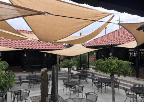 Outdoor dining area with shaded tables, decorative lights, and a rustic building in the background.