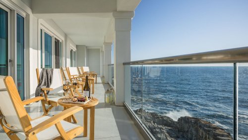 A seaside balcony with wooden chairs and tables overlooking the ocean, featuring clear blue skies and calm waters.