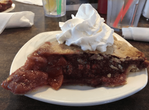 A slice of pie with a flaky crust and cherry filling, topped with a generous dollop of whipped cream on a white plate.