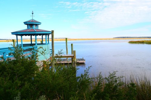 A blue gazebo by a calm river, surrounded by greenery and a clear sky, with marshland in the background.