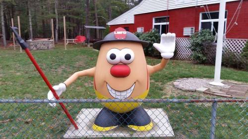 A large, cartoonish potato figure wearing a firefighter hat, waving and holding a red pole, in a fenced yard.