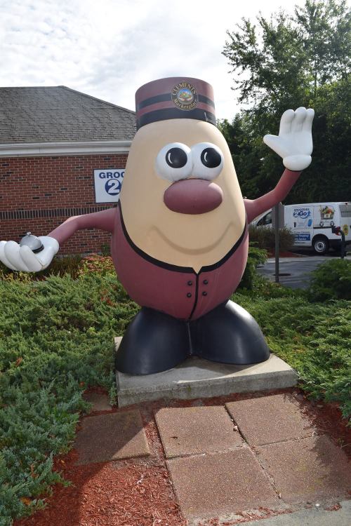 A large, cartoonish potato mascot in a bellhop uniform, waving outside a grocery store.